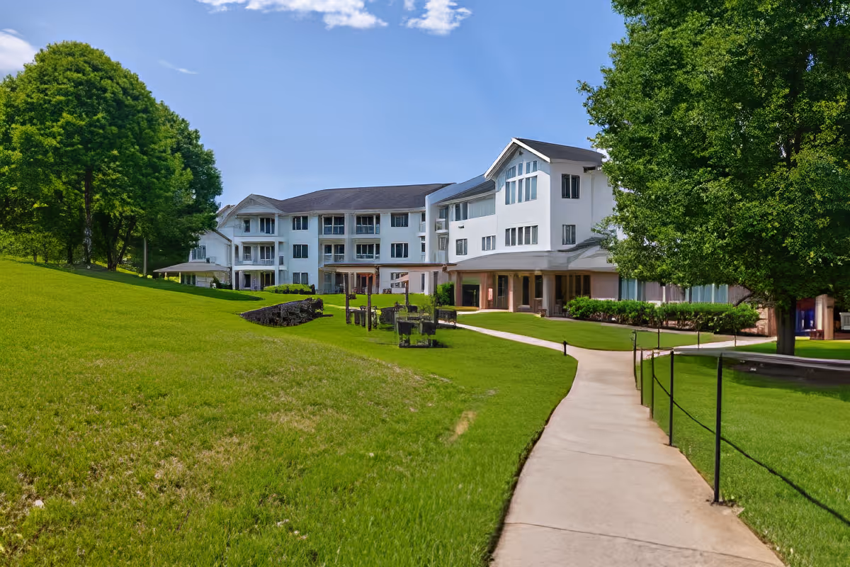 Front exterior of a multi-story senior living building with a paved walkway, green lawn, and trees.