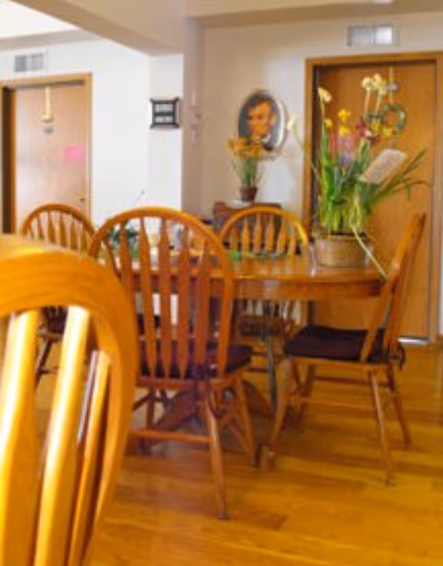 Wooden dining table with matching chairs and a floral centerpiece in a communal dining room with doors and a portrait on the wall.