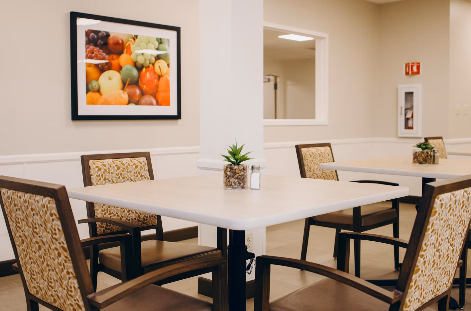 A dining area with square tables and cushioned chairs featuring a patterned fabric. Each table has a small plant and a salt shaker. The walls are beige with white trim, and there is a framed picture of assorted fruits hanging on the wall. A fire extinguisher is visible in the background.