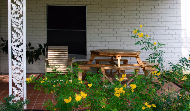 Outdoor patio area with a wooden picnic table and benches, a wooden chair, and yellow flowering plants in front. The background features a white brick wall with a window and a decorative white metal post.
