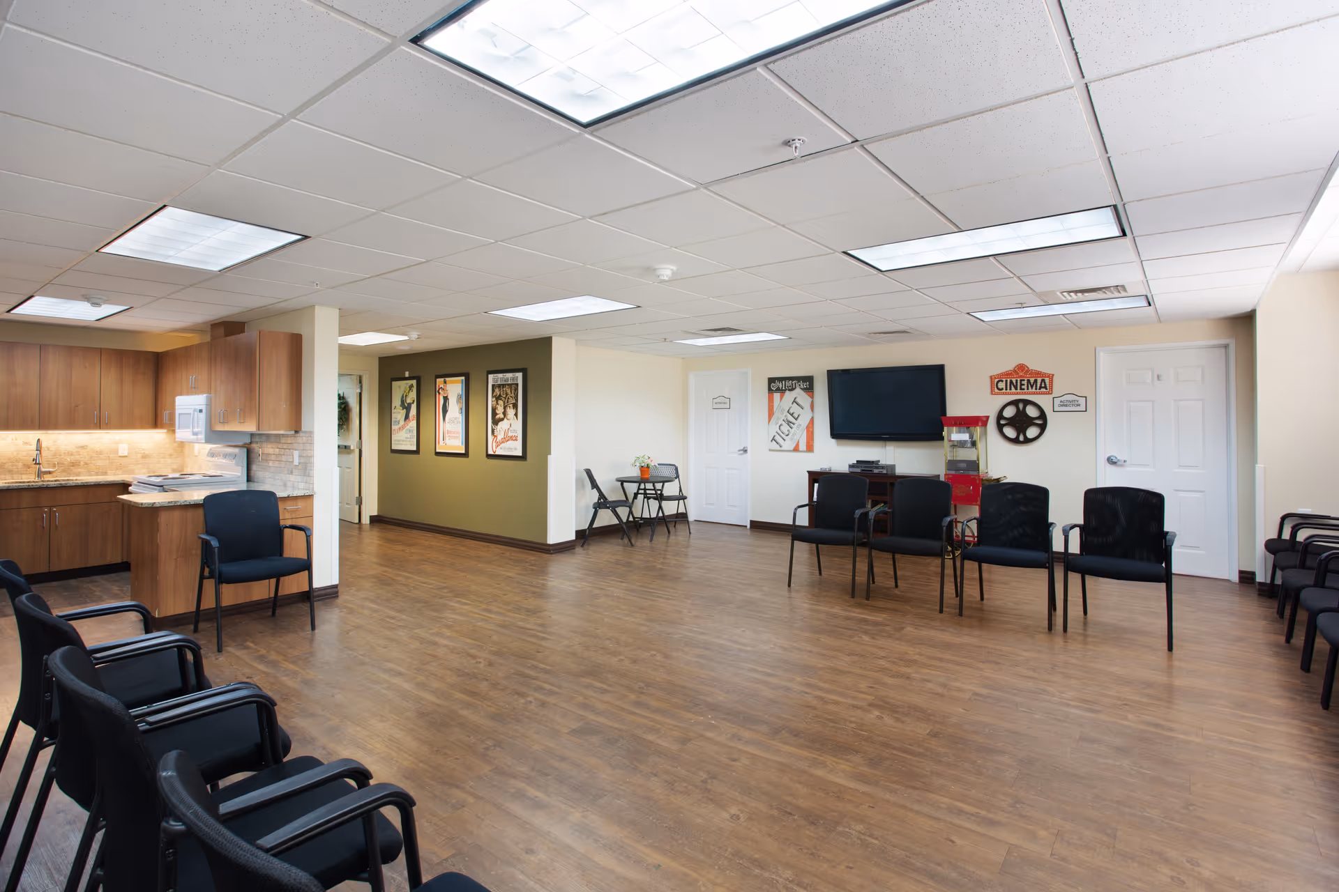 A spacious common room with wood flooring and a drop ceiling with fluorescent lights. The room features a small kitchenette with wooden cabinets, a microwave, and a sink on the left side. Several black chairs are arranged along the walls and in the center of the room. On the far wall, there is a flat-screen TV mounted above a cabinet, a red popcorn machine, and movie-themed decorations including a cinema sign, a film reel, and vintage movie posters. Two white doors are visible on the right side of the room.