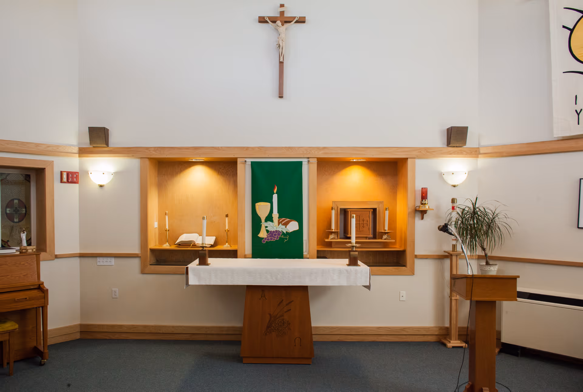 Chapel interior showing an altar with a white cloth, crucifix on the wall, candles, a green liturgical banner, and a lectern.