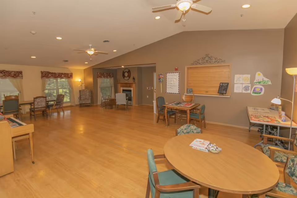 A spacious common room in McFarland Villa Assisted Living with wooden floors, several tables and chairs arranged for group activities, a fireplace in the background, ceiling fans, and windows with curtains letting in natural light. The walls have some decorations and artwork, and there is a piano on the left side.