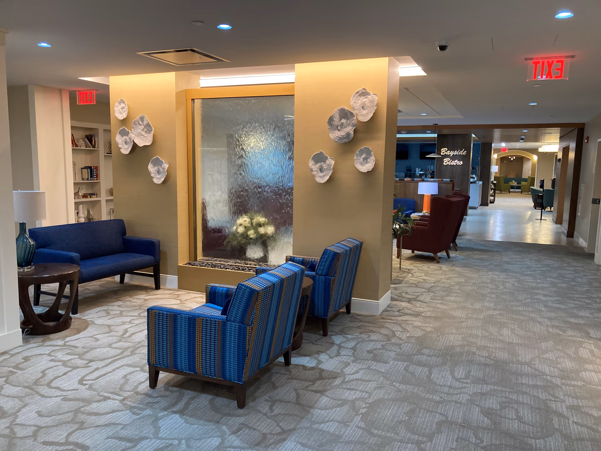 Lobby lounge with blue upholstered chairs facing a decorative water wall and a view down the corridor to the Bayside Bistro.