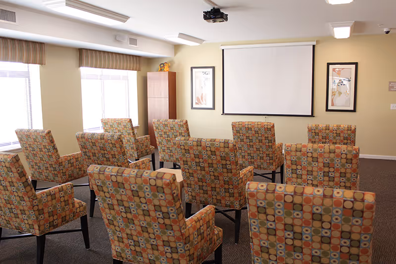 Small meeting/screening room with patterned armchairs arranged facing a wall-mounted projection screen and projector.