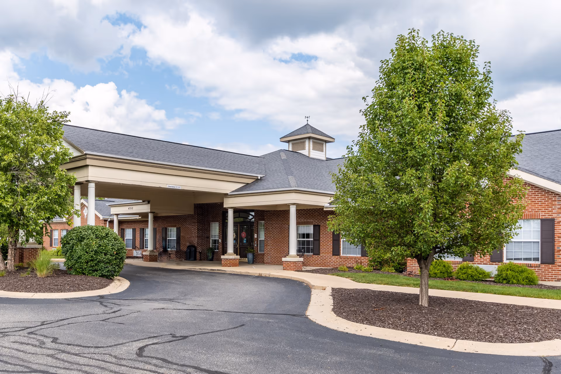 Exterior view of Walnut Ridge Assisted Living and Memory Care building with a covered entrance, brick walls, windows with shutters, and landscaped greenery including trees and bushes under a partly cloudy sky.
