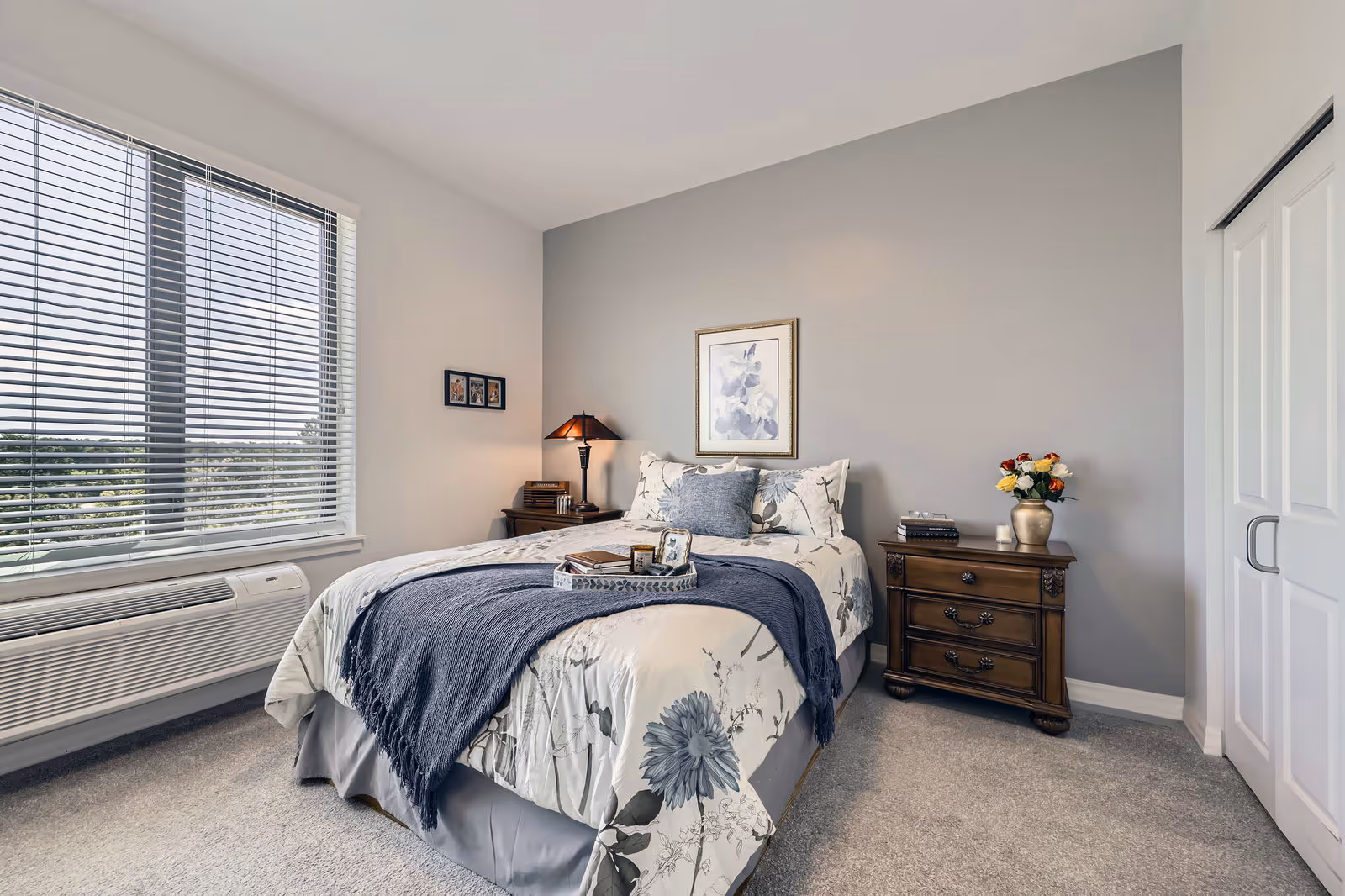A neatly made bed with floral bedding and a dark blue throw blanket in a bright bedroom. The room has a large window with blinds, a wooden nightstand with a lamp and books on one side, and another wooden nightstand with a vase of flowers and a candle on the other side. There is a framed floral artwork on the light gray wall above the bed and a closet with white sliding doors on the right.