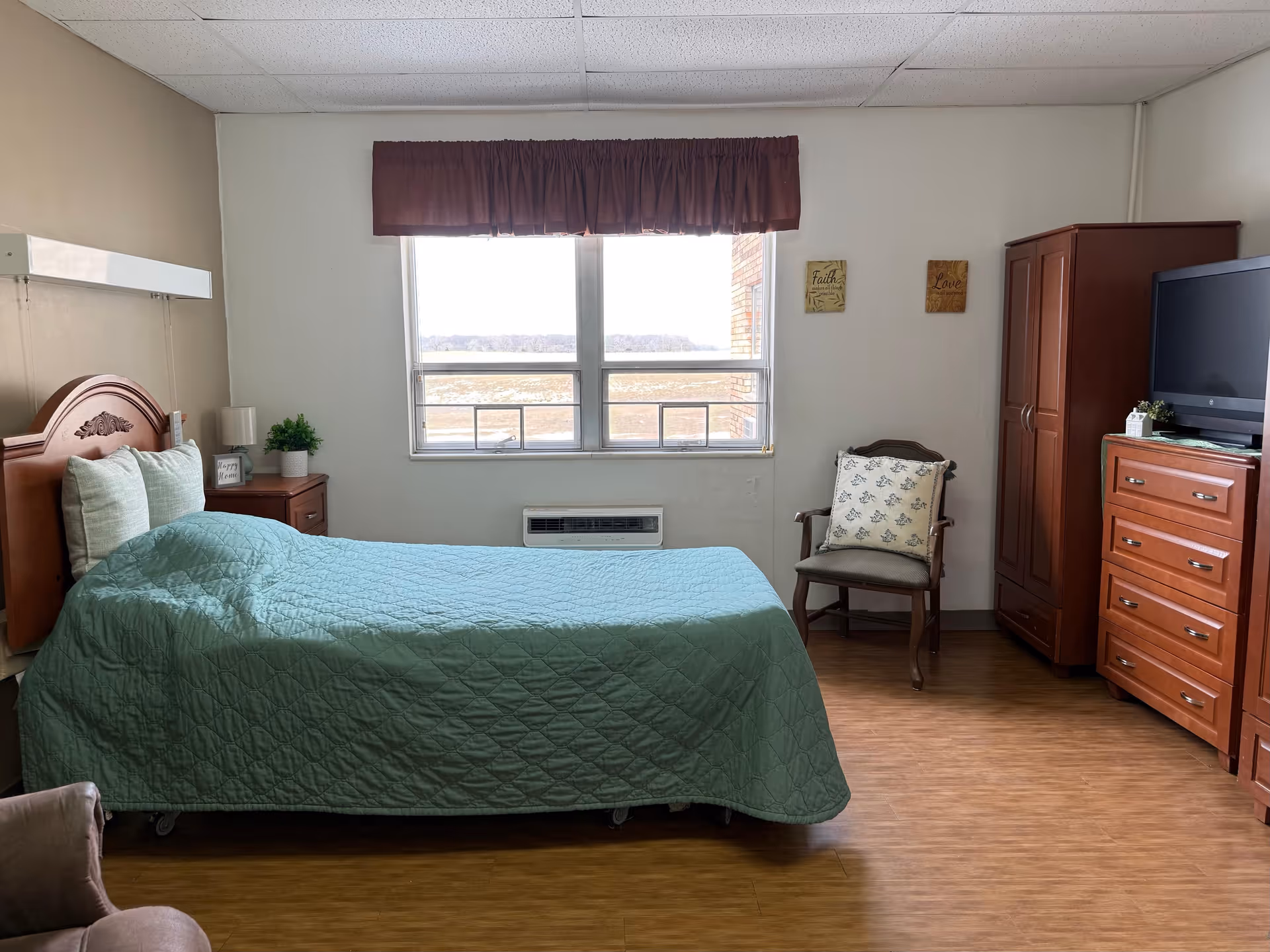 A neatly arranged bedroom in a senior living facility with a single bed covered in a green quilt, two pillows, a wooden headboard, a nightstand with a lamp and a small plant, a window with a maroon valance, a wooden chair with a cushion, a wooden wardrobe, a dresser with a TV on top, and two small wall decorations.