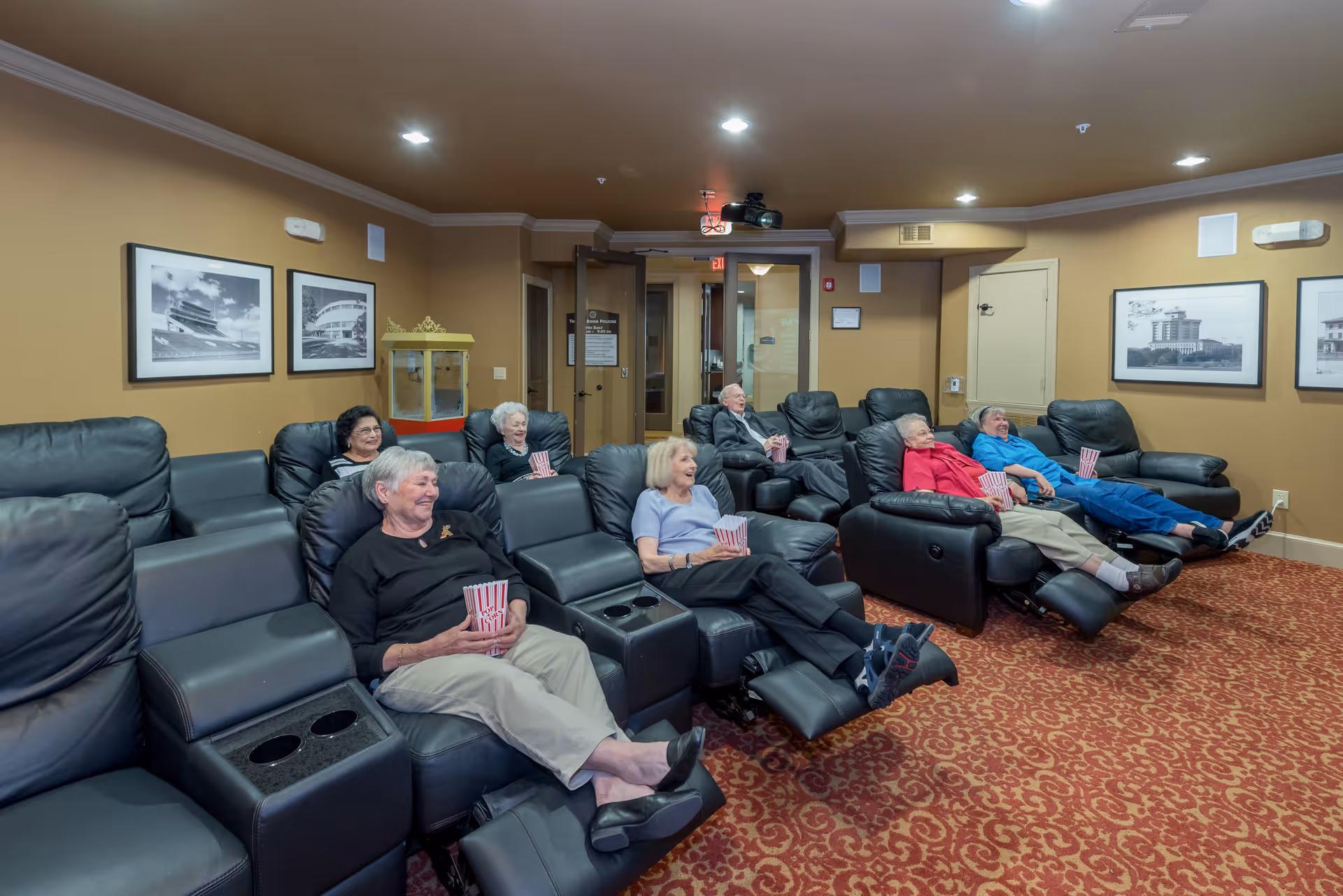 A group of elderly people sitting in black leather recliners in a cozy movie theater room, each holding a container of popcorn. The room has warm beige walls, framed black and white photos, and a red patterned carpet.