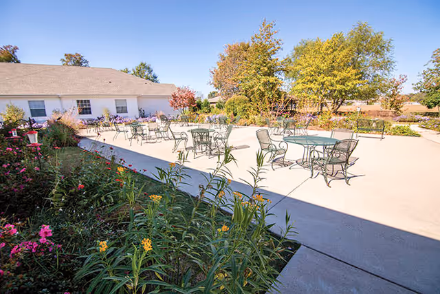 Outdoor patio area with several metal tables and chairs arranged on a concrete surface, surrounded by colorful flowers and greenery, with a white building and trees in the background under a clear blue sky.