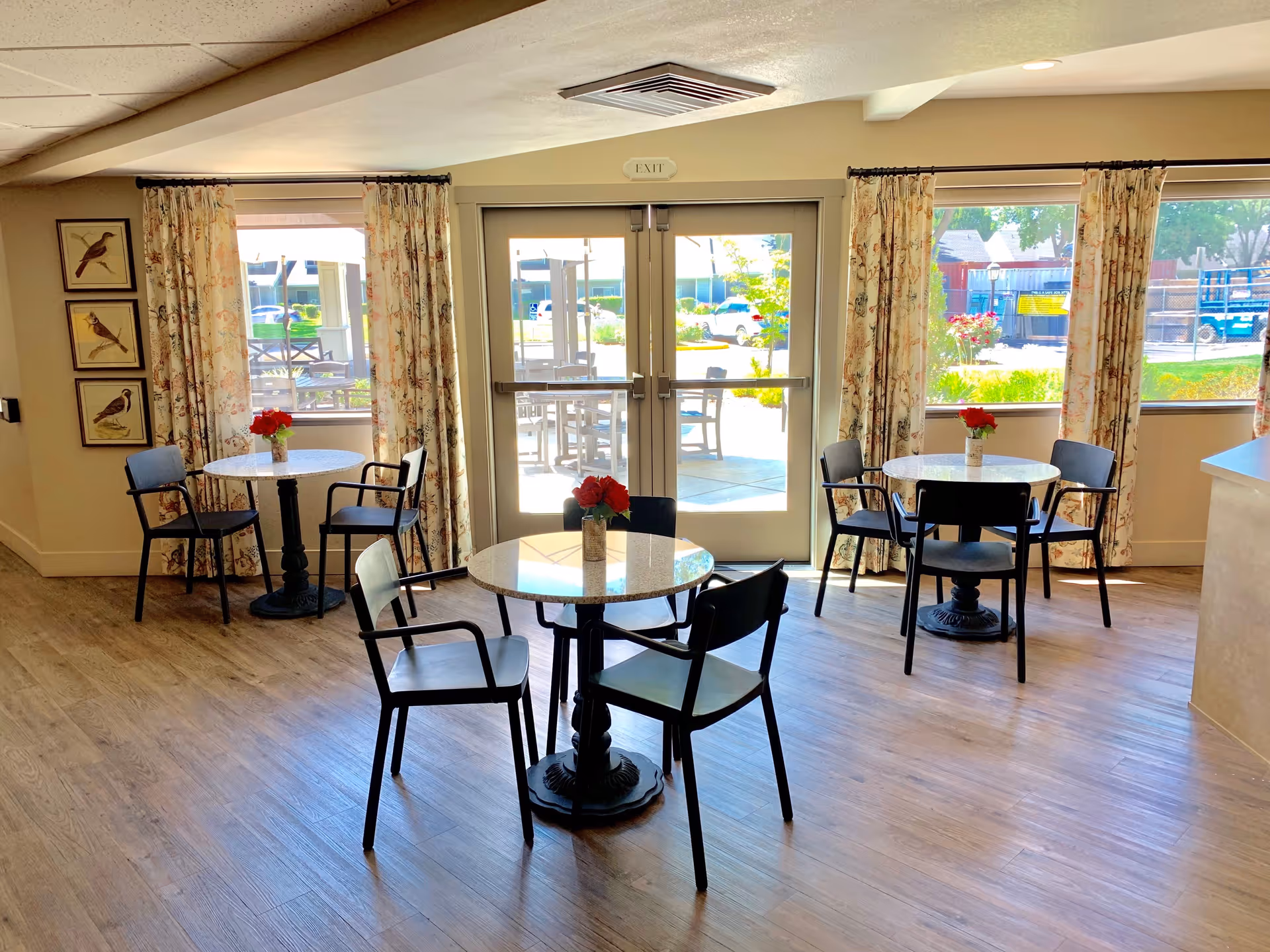 A bright dining area with three round tables, each surrounded by black chairs. Each table has a small vase with red flowers. Large windows with floral curtains let in natural light, and a glass door leads outside to a patio area with more tables and chairs. The floor is wooden, and there are framed bird pictures on the wall to the left.