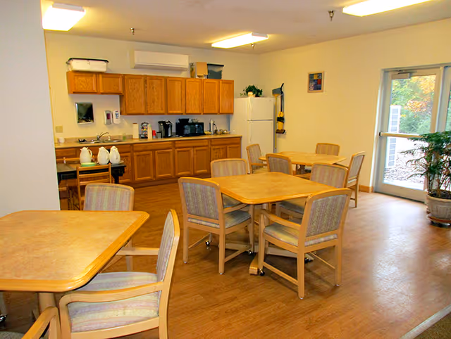 A dining area with several wooden tables and chairs arranged on a wood floor. In the background, there is a kitchenette with wooden cabinets, a refrigerator, a microwave, and a coffee maker. A door with a window leads outside, and a potted plant is near the door.