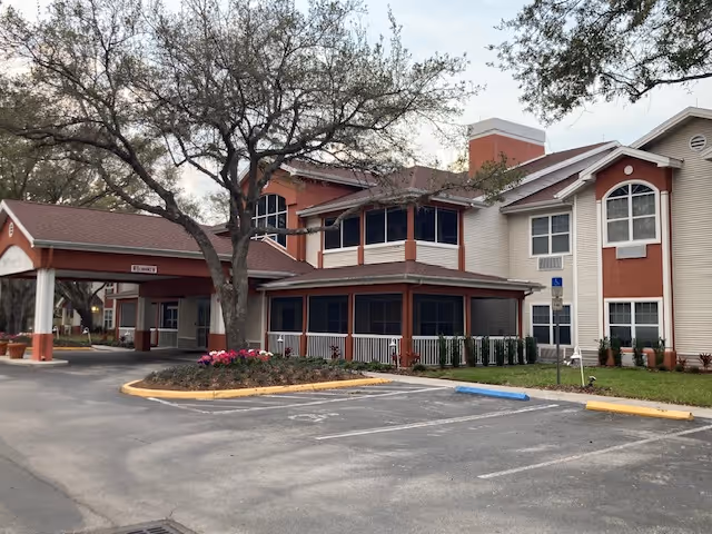 Exterior view of a two-story senior living facility building with beige and reddish-brown walls, large windows, a covered entrance, and surrounding trees and landscaping. There is a parking lot with marked spaces in front of the building.