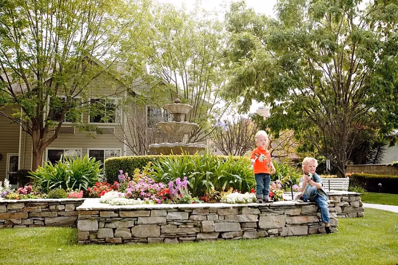 Two young children playing near a stone flower bed with colorful flowers and a tiered water fountain in a green outdoor garden area with trees and a building in the background.