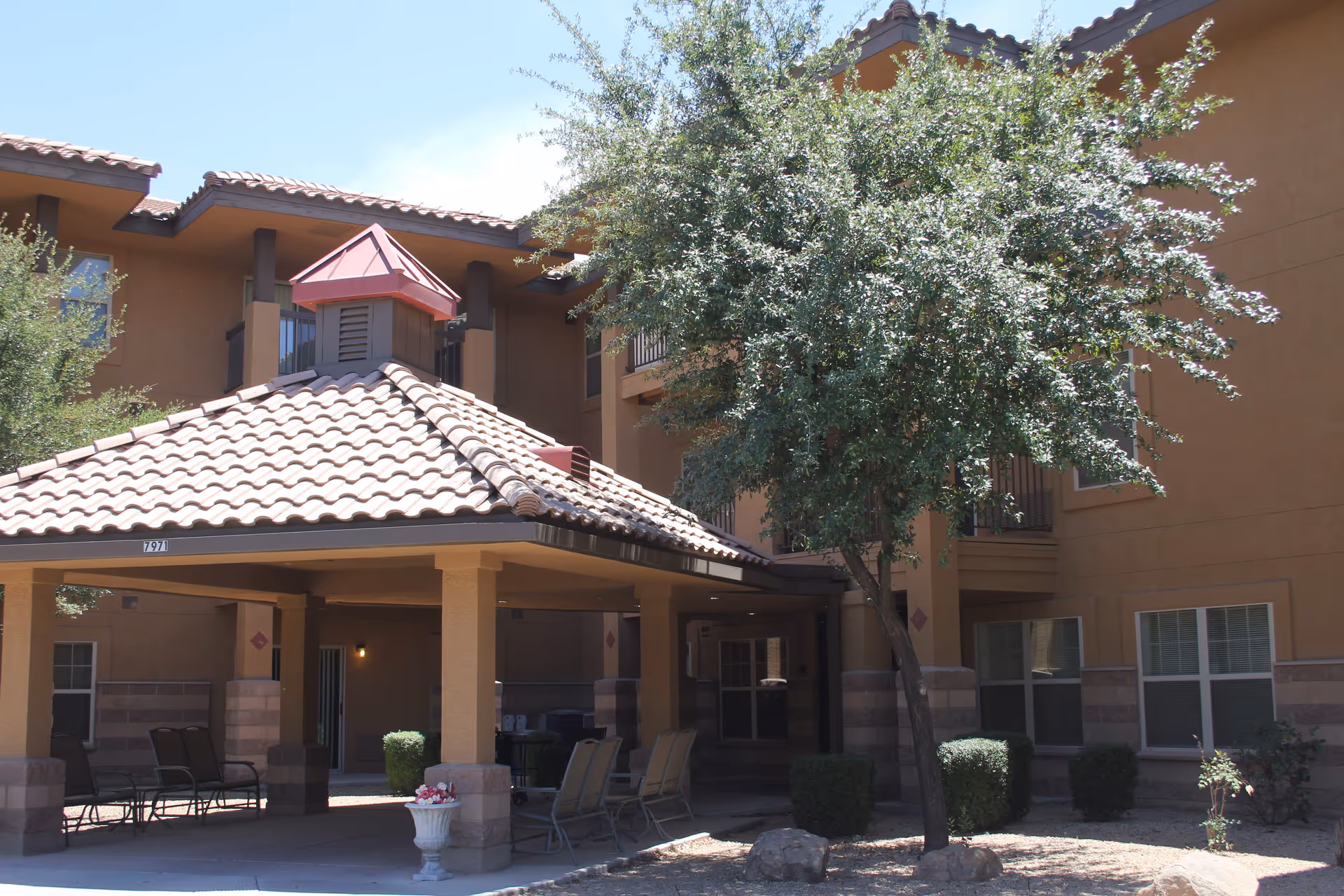 Exterior view of a senior living facility building with a covered patio area featuring several chairs and a small table. The building has a tiled roof and beige walls with multiple windows. There is a tree and some bushes in the landscaped area near the patio.