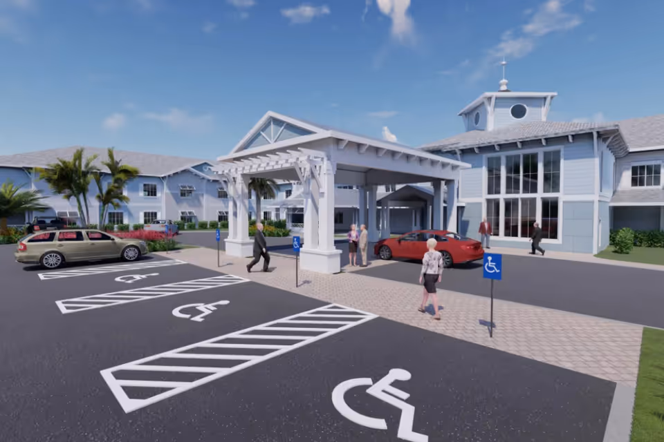 Exterior view of The Meridian at Punta Gorda Isles senior living facility showing the entrance with a covered drop-off area, several parked cars, accessible parking spaces, palm trees, and people walking near the entrance on a sunny day.