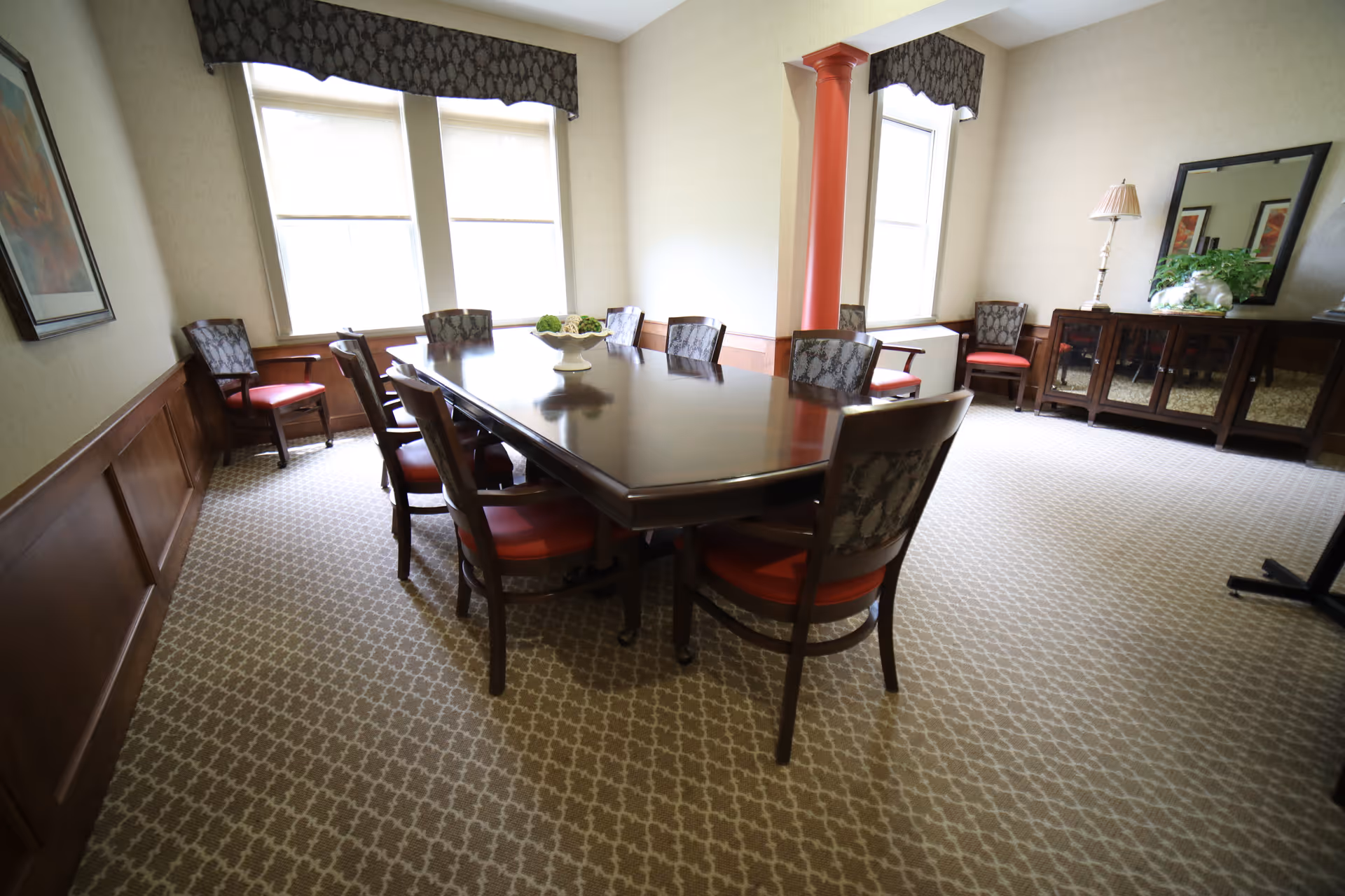 A formal dining room with a large polished wooden table surrounded by chairs, windows, and a sideboard.