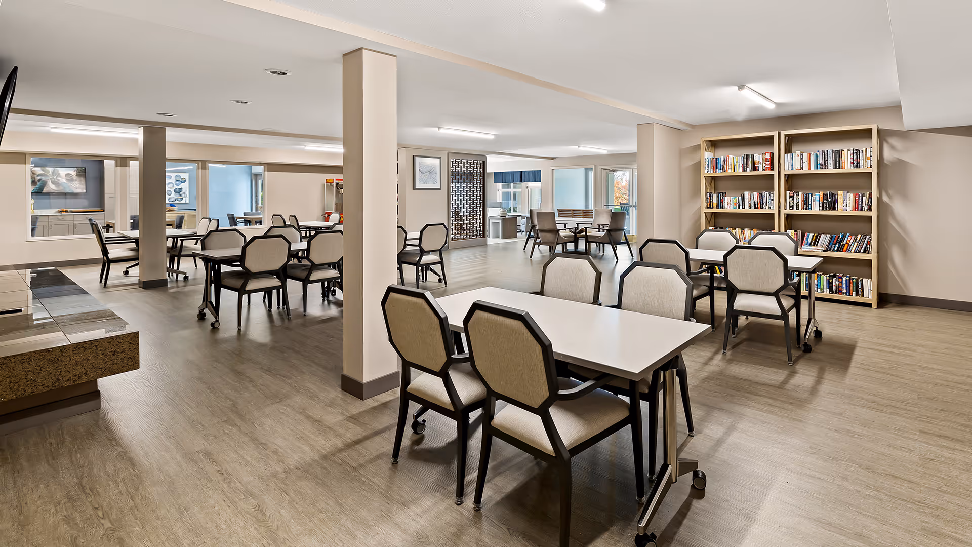 A spacious, well-lit common area with multiple tables and chairs arranged for group seating. There are bookshelves filled with books along one wall, and large windows allowing natural light to enter. The room has a clean, modern design with light-colored walls and wood flooring.