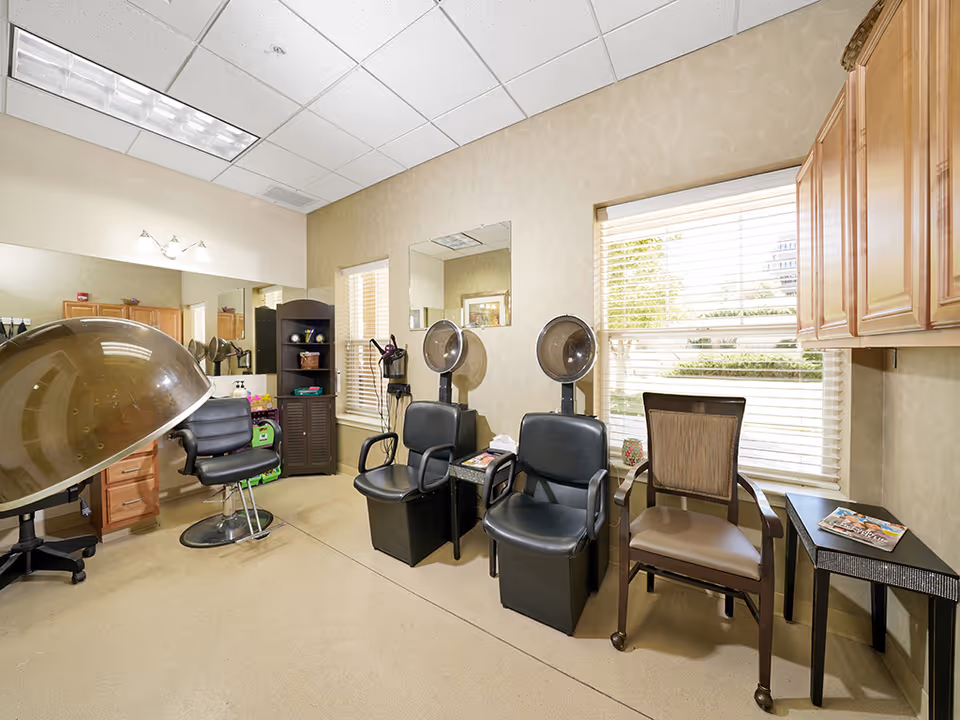 Interior of a hair salon area in a senior living facility with two black salon chairs under hair dryers, a black styling chair, a wooden chair, a small table with magazines, a tall black shelving unit, and wooden cabinets. Large windows with blinds allow natural light to fill the room.