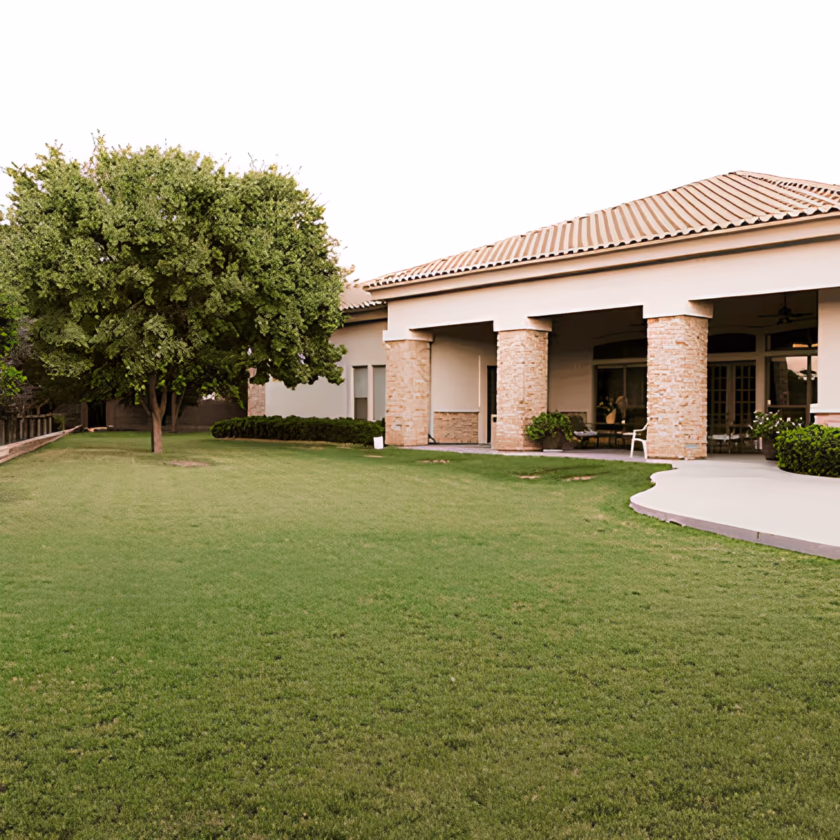 A large green lawn with a single leafy tree on the left side and a building with a tiled roof and stone pillars on the right side, featuring a covered patio area with seating.