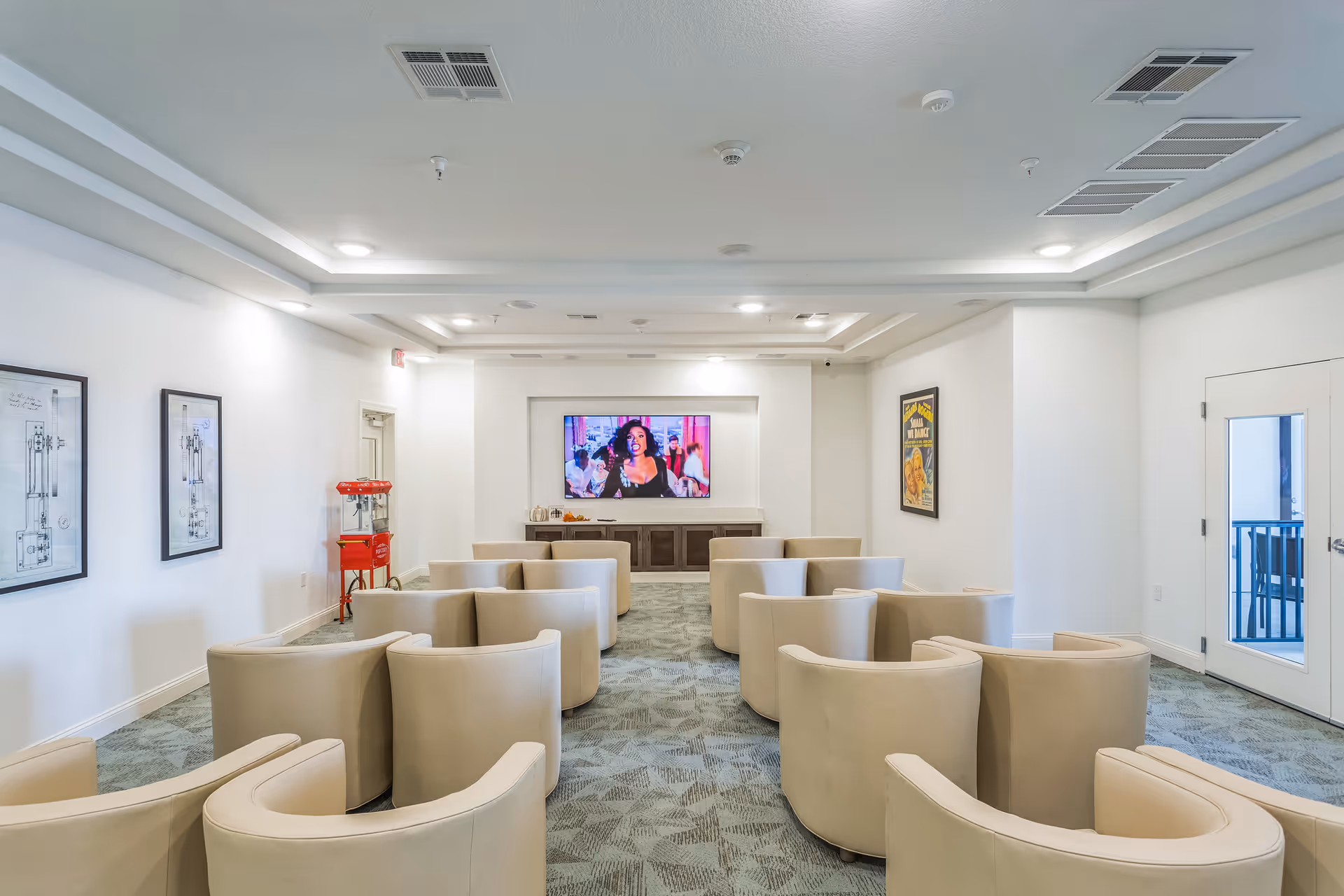 A bright media room with curved beige chairs arranged in rows facing a wall-mounted television and a popcorn machine.