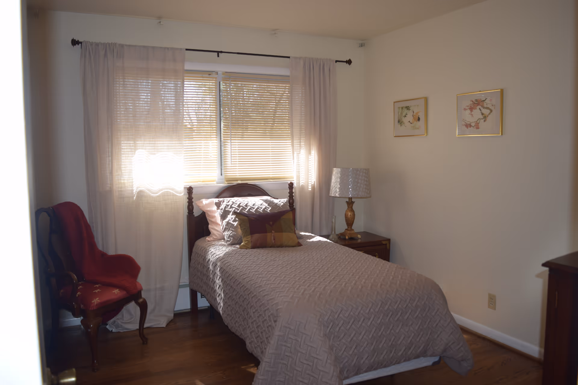 A cozy bedroom with a single bed covered in a textured beige bedspread and multiple pillows. A wooden chair with a red cushion and a red throw blanket is placed near the window, which has sheer curtains and blinds. A wooden nightstand with a decorative lamp and a small glass vase is beside the bed. Two framed pictures hang on the light-colored wall.