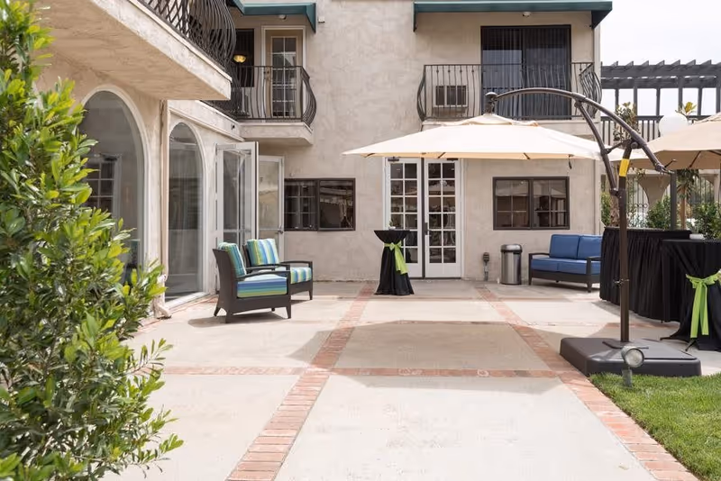 Outdoor patio area at Sunnycrest Senior Living with cushioned chairs, a large umbrella, a small table with a black cover and green ribbon, and a two-story building with balconies and windows in the background.
