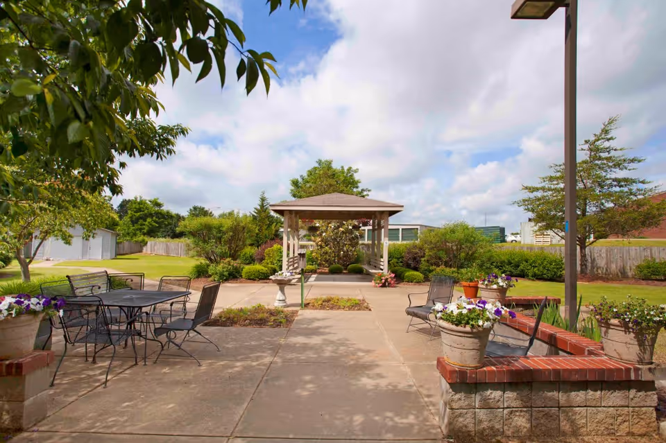 Outdoor patio area with metal tables and chairs, surrounded by flower pots with blooming flowers. A paved walkway leads to a wooden gazebo in the background, with green bushes, trees, and a partly cloudy sky.