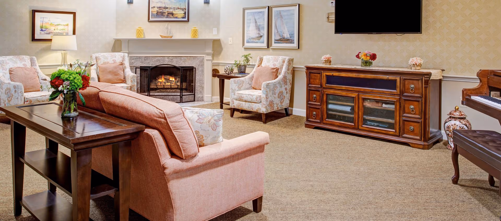 Comfortable living room with upholstered sofa and chairs arranged around a lit fireplace, a TV mounted above a wooden sideboard, and floral decor.