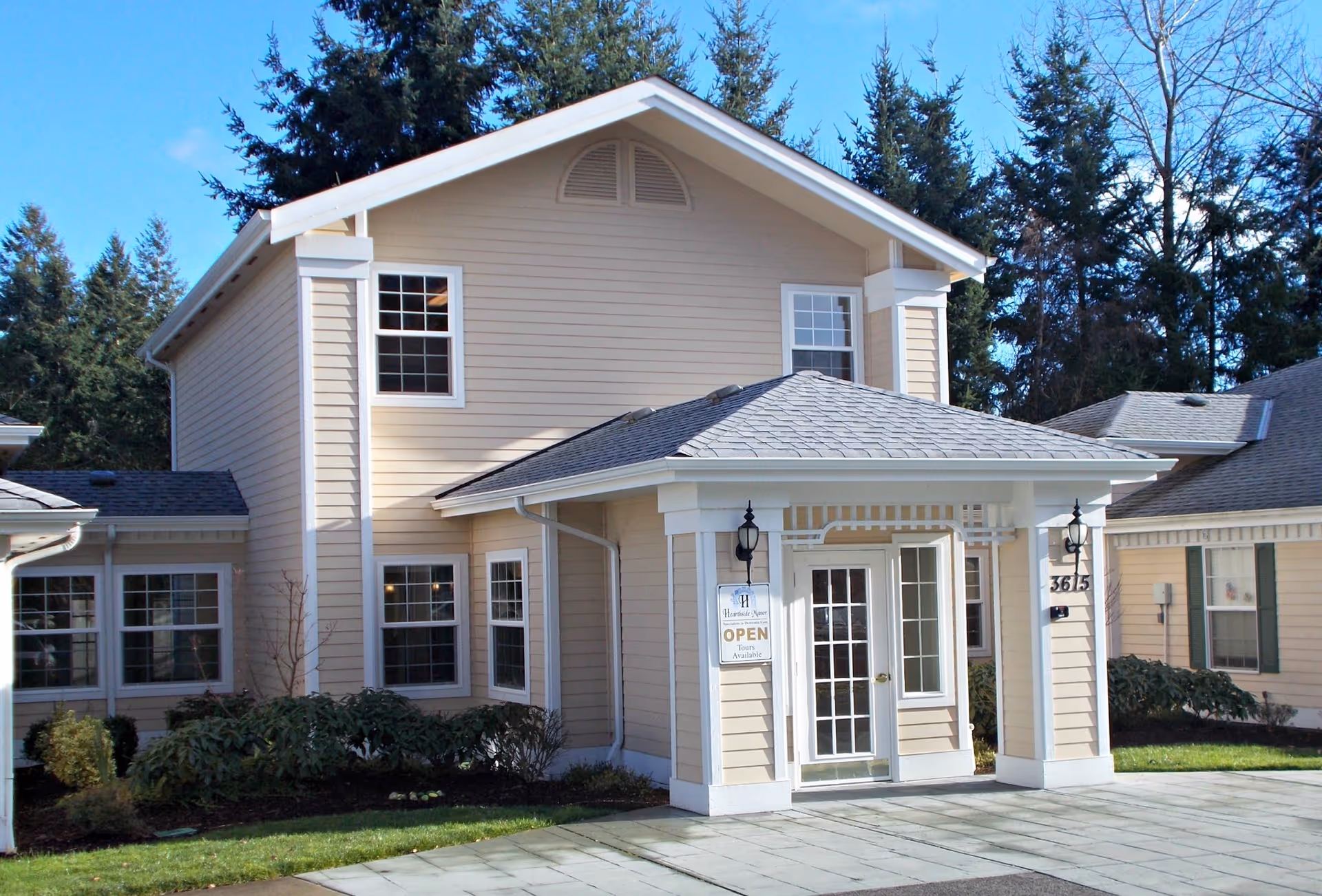 Exterior view of a beige two-story senior living facility building with white trim, a covered entrance with glass double doors, and a sign indicating it is open for tours. The building is surrounded by greenery and tall trees in the background under a clear blue sky.