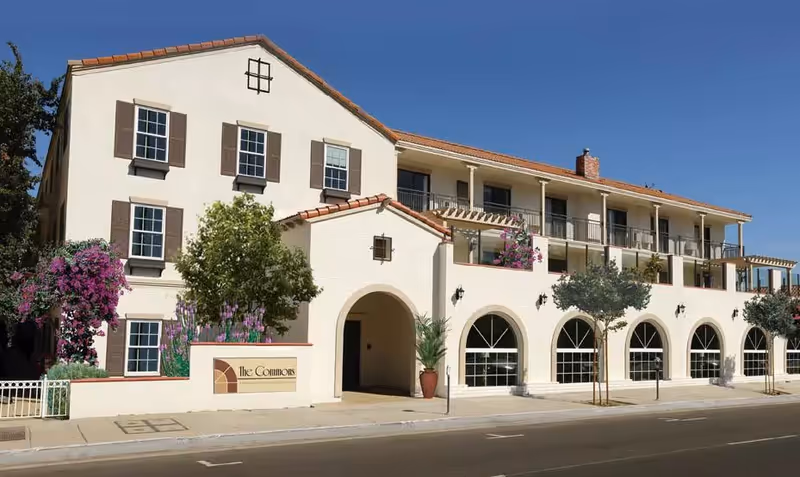 Exterior view of a multi-story senior living facility building with beige walls, brown window shutters, and a red-tiled roof under a clear blue sky. The building has arched windows on the ground floor and balconies on the upper floor, with some potted plants and small trees along the sidewalk. A sign near the entrance reads 'The Commons'.