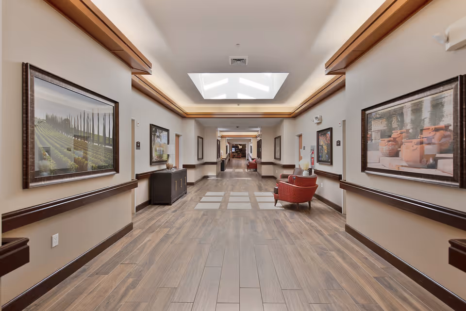 A well-lit hallway in a senior living facility with wooden flooring, beige walls, framed landscape artwork on both sides, and red upholstered chairs placed along the walls. The ceiling features recessed lighting and a skylight.