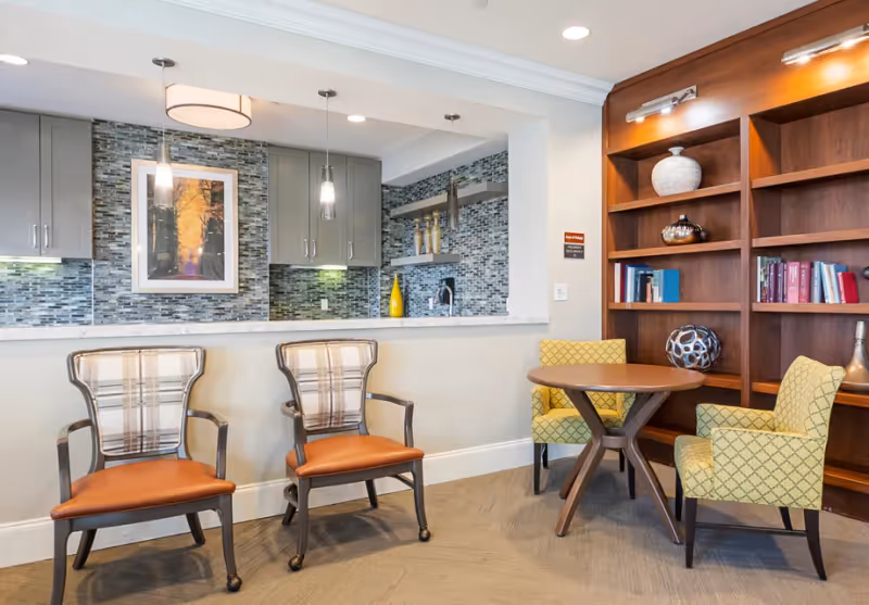 A furnished common area with two plaid-back armchairs at a counter, a small round table with two patterned chairs, and a wooden bookshelf.