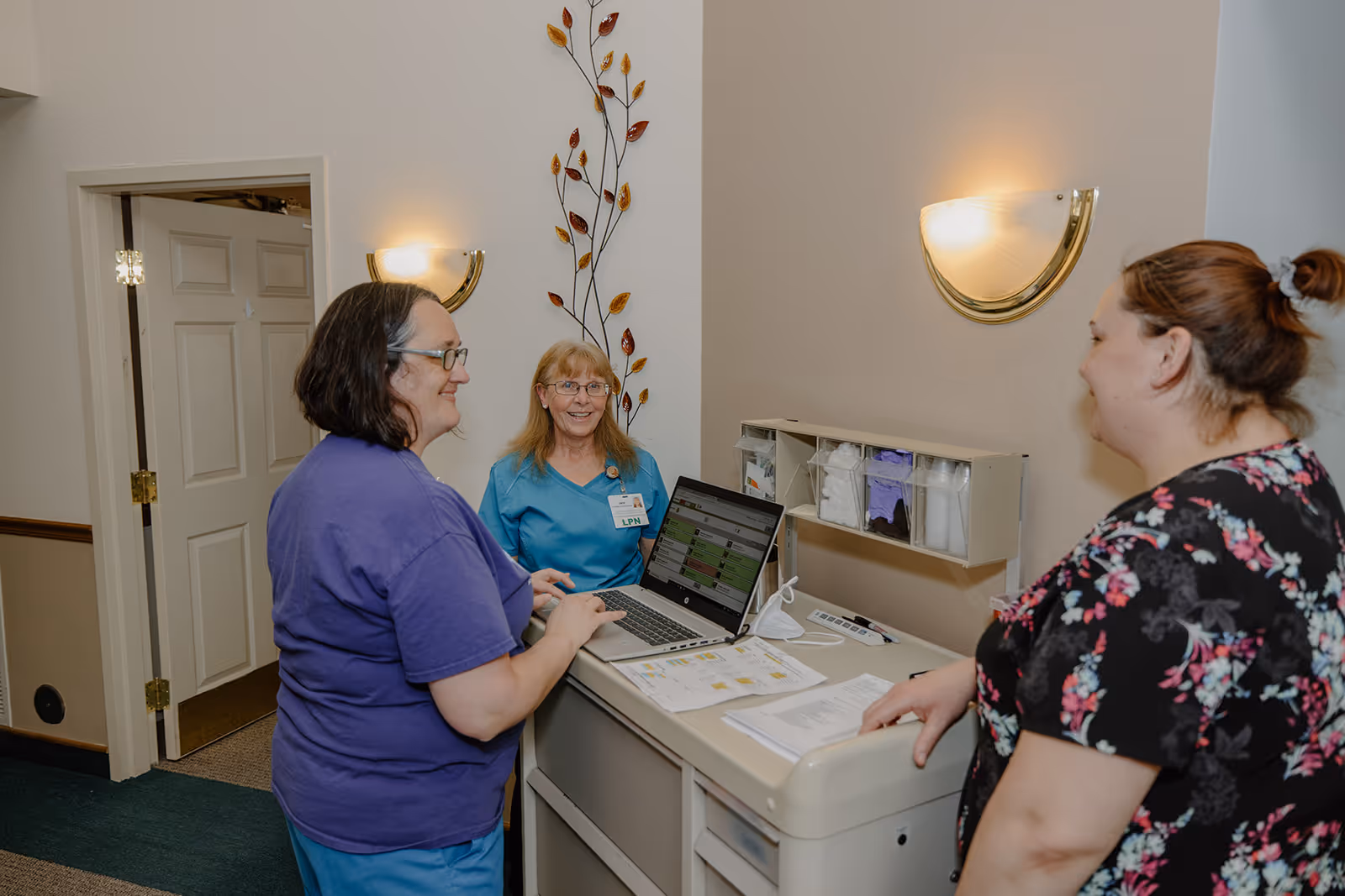 Three women standing and talking around a medical cart with a laptop and papers in a hallway of an assisted living facility. One woman is wearing a blue medical uniform with an LPN badge, another is in a purple shirt, and the third is in a black floral top. The hallway has beige walls with decorative wall sconces and a metal wall decoration with leaves.