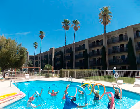 Outdoor swimming pool with several people participating in a water exercise class using colorful pool noodles. The pool is surrounded by a white fence, lounge chairs, and tall palm trees. In the background, there is a multi-story residential building under a clear blue sky.