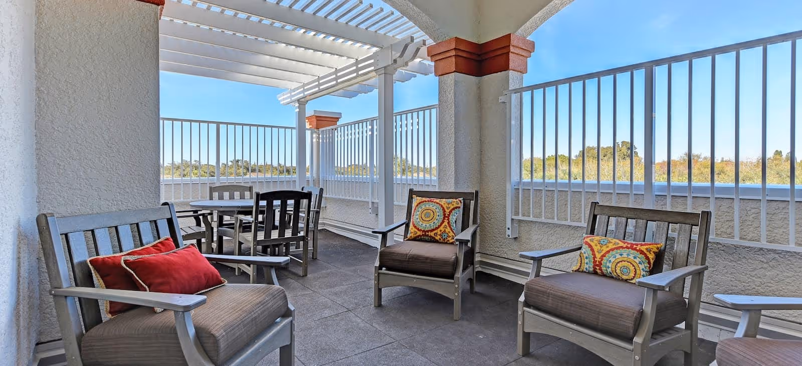 Outdoor seating area with cushioned wooden chairs and a round table under a white pergola with a view of trees and clear blue sky.