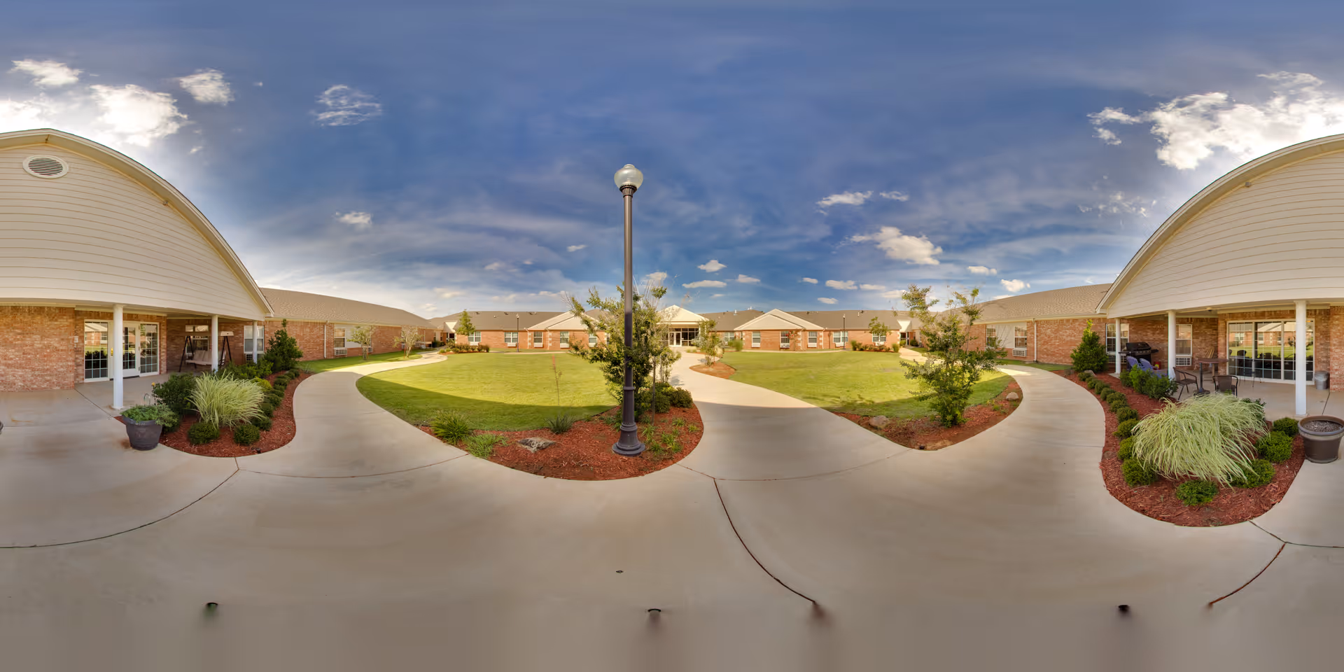 Wide panoramic view of an outdoor courtyard at Plantation Village Nursing Center featuring a curved concrete walkway, green lawn, small trees, shrubs, and two covered porch areas with seating. The sky is partly cloudy with blue tones.