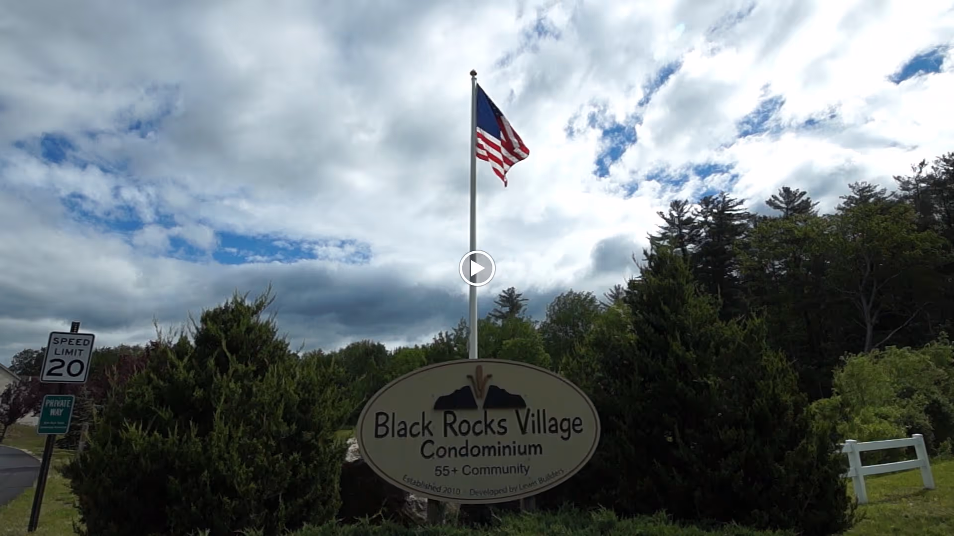 Entrance sign for Black Rocks Village Condominium, a 55+ community, with an American flag flying on a flagpole above it and trees in the background under a partly cloudy sky.