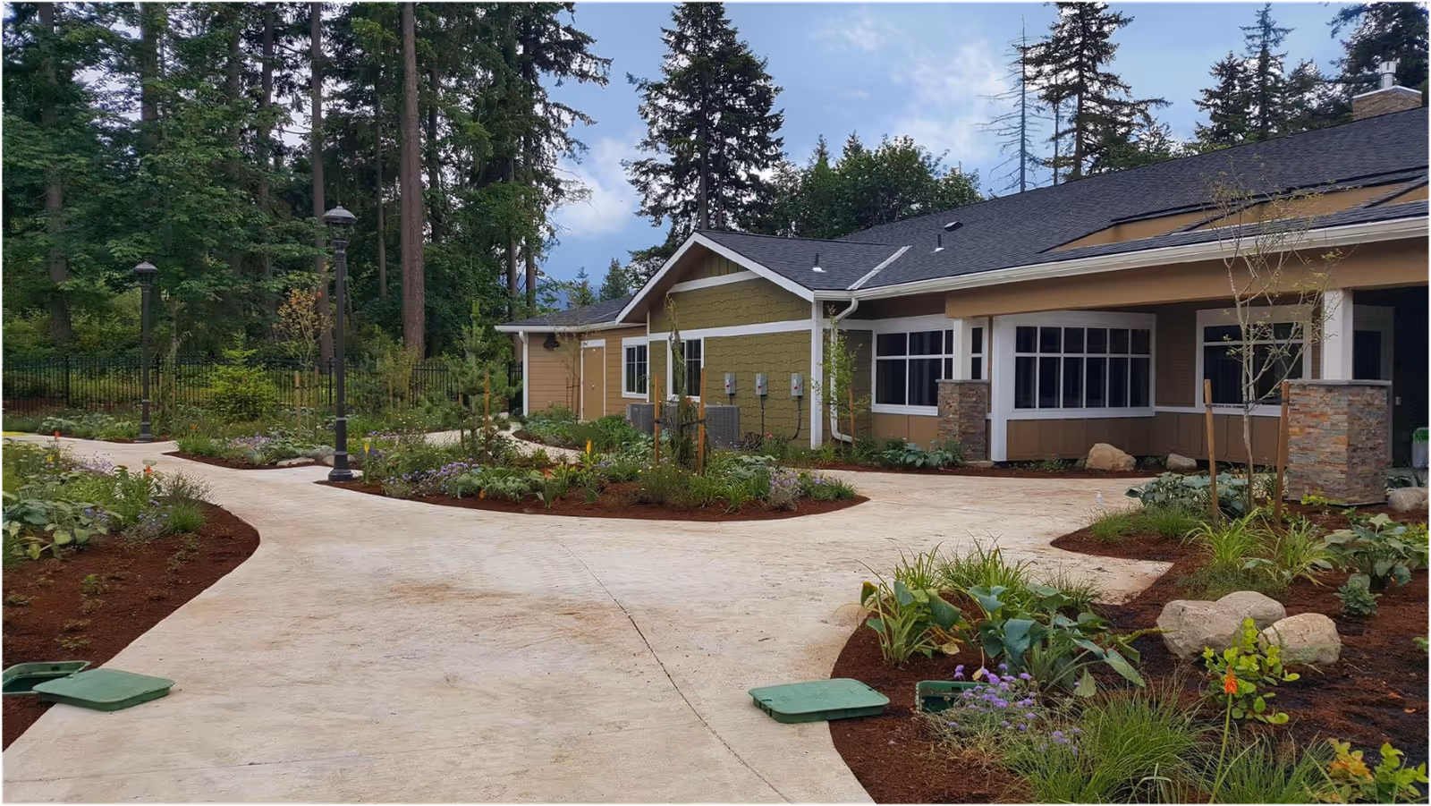 Outdoor view of a senior living facility with a paved walkway surrounded by landscaped garden beds, plants, and trees. The building has large windows and a sloped roof with solar panels, set against a backdrop of tall trees and a partly cloudy sky.