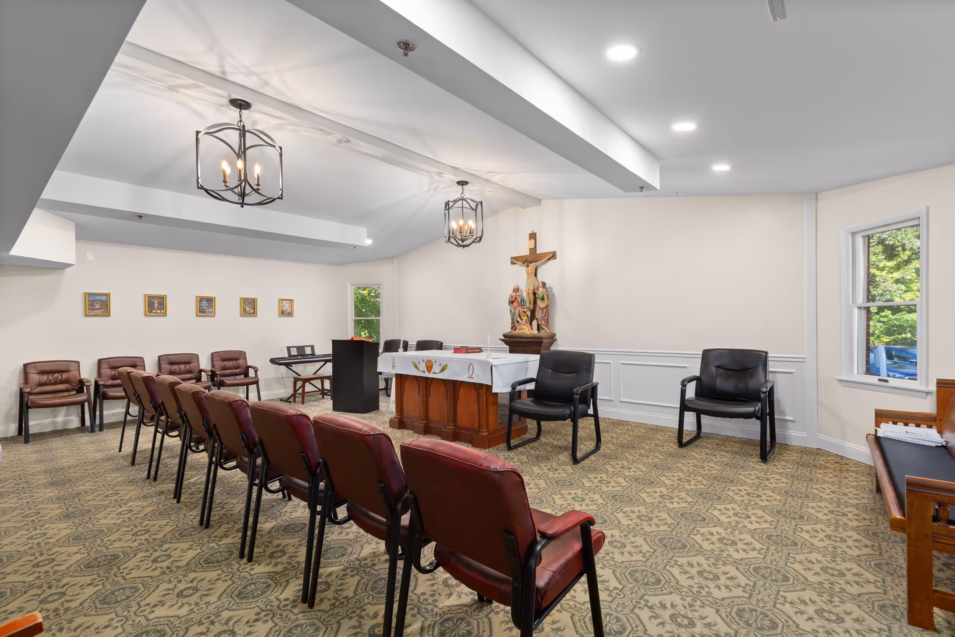 A small chapel or prayer room with rows of brown chairs facing a wooden altar covered with a white cloth. Behind the altar is a crucifix with religious statues. The room has patterned carpet, white walls, two windows, and two black chandeliers hanging from the ceiling.