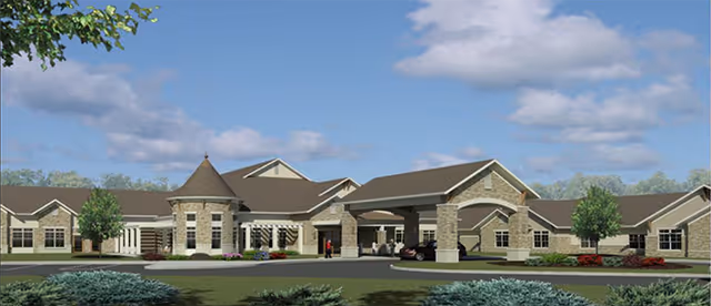 Exterior view of The Gables of Green senior living facility showing a large, single-story building with a covered entrance, stone and beige siding, multiple windows, and a turret-like architectural feature under a partly cloudy sky.