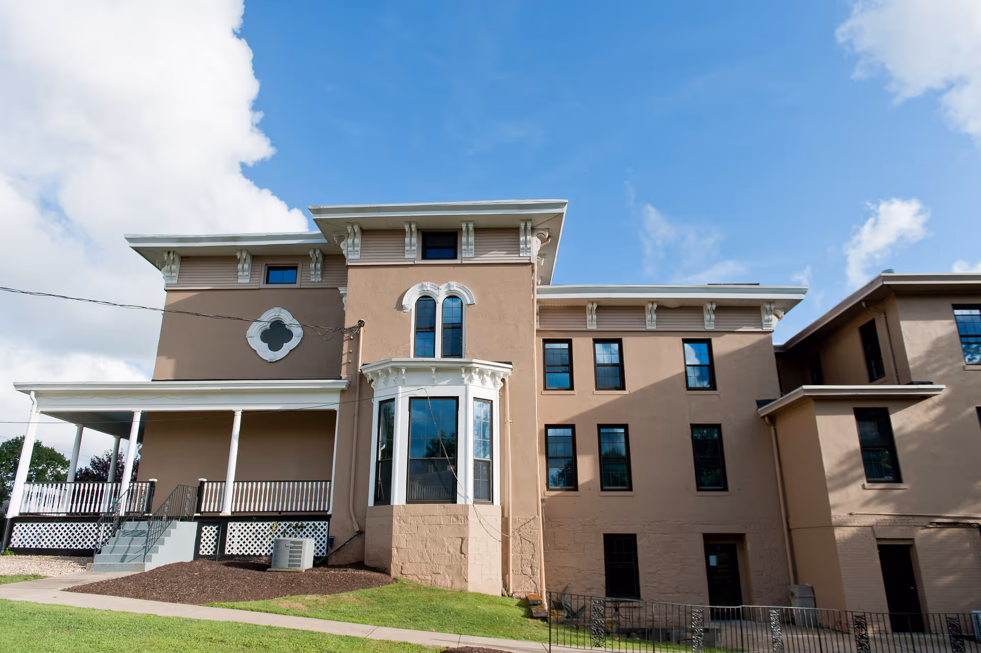 Exterior view of a multi-story beige building with white decorative trim and multiple windows under a blue sky with some clouds. The building has a covered porch with white columns and a small lawn in front.