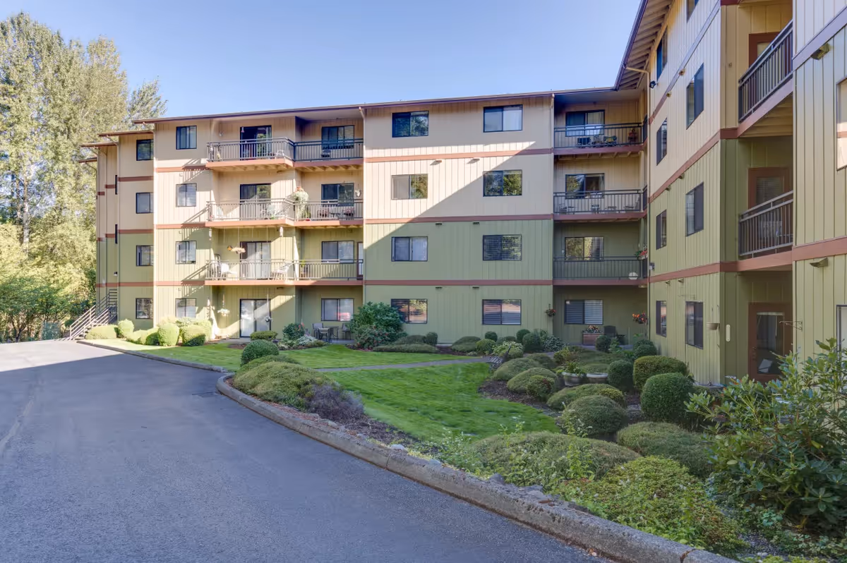 Exterior view of a multi-story residential building with balconies, surrounded by well-maintained green lawns and shrubs under a clear blue sky.