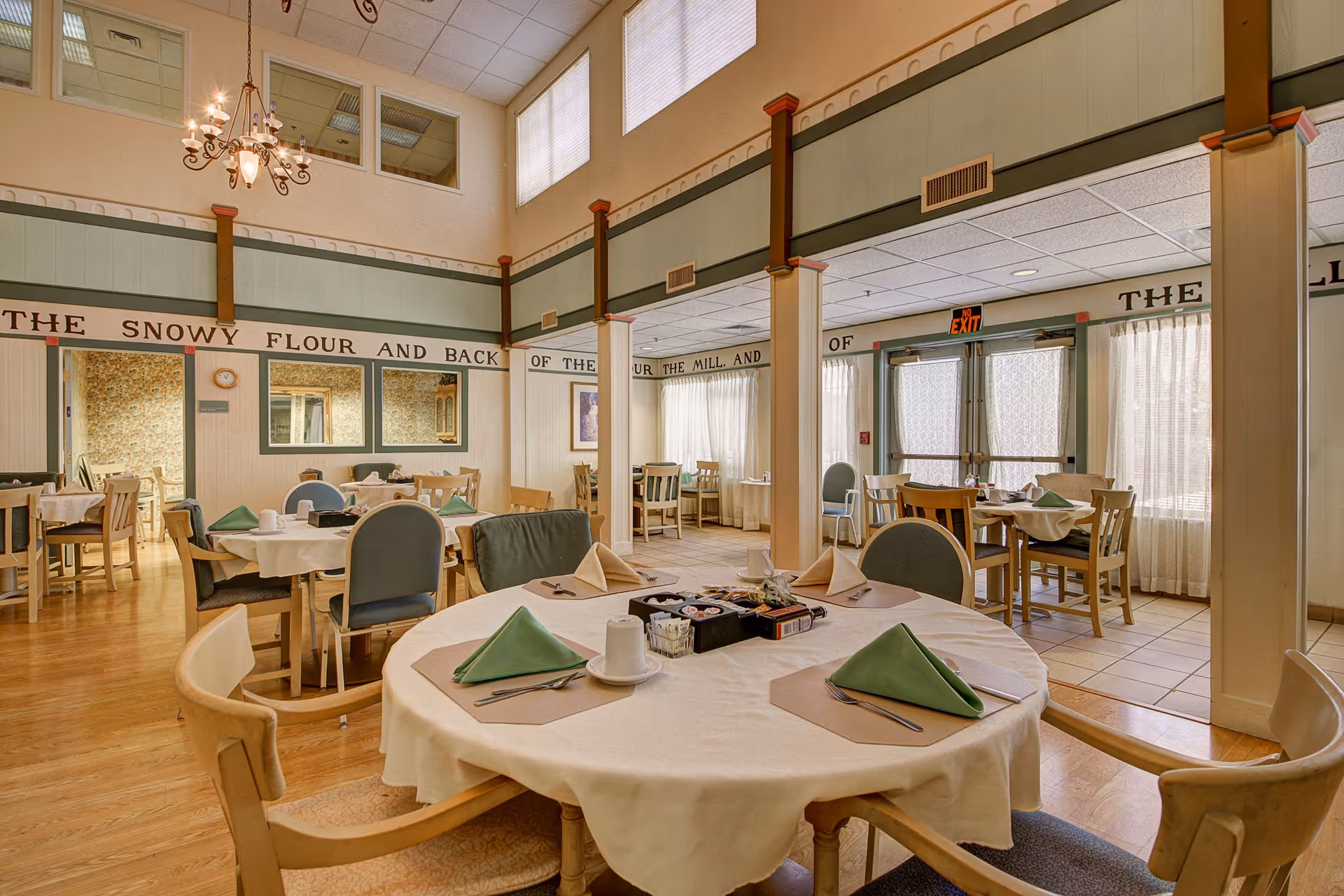 A bright dining room in an assisted living facility with round and square tables set with white tablecloths, green folded napkins, cups, and utensils. The room has wooden floors, large windows with sheer curtains, and a chandelier hanging from the ceiling. There are multiple chairs around each table and a decorative wall with text running along the top.