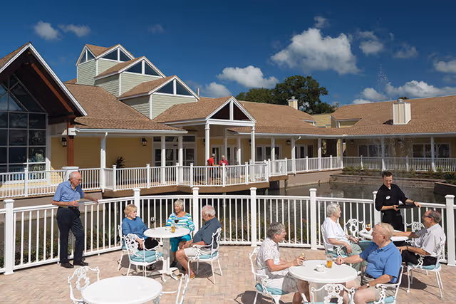 Elderly residents sit at white patio tables while a staff member serves them on an outdoor terrace by a pond in front of a yellow building.
