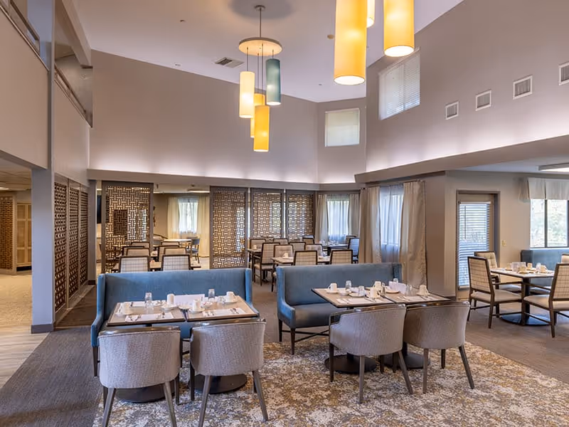 A spacious dining room with multiple tables set for meals, featuring cushioned chairs and booth seating. The room has high ceilings with modern hanging pendant lights in various colors, large windows with curtains, and decorative wooden partitions separating different seating areas.
