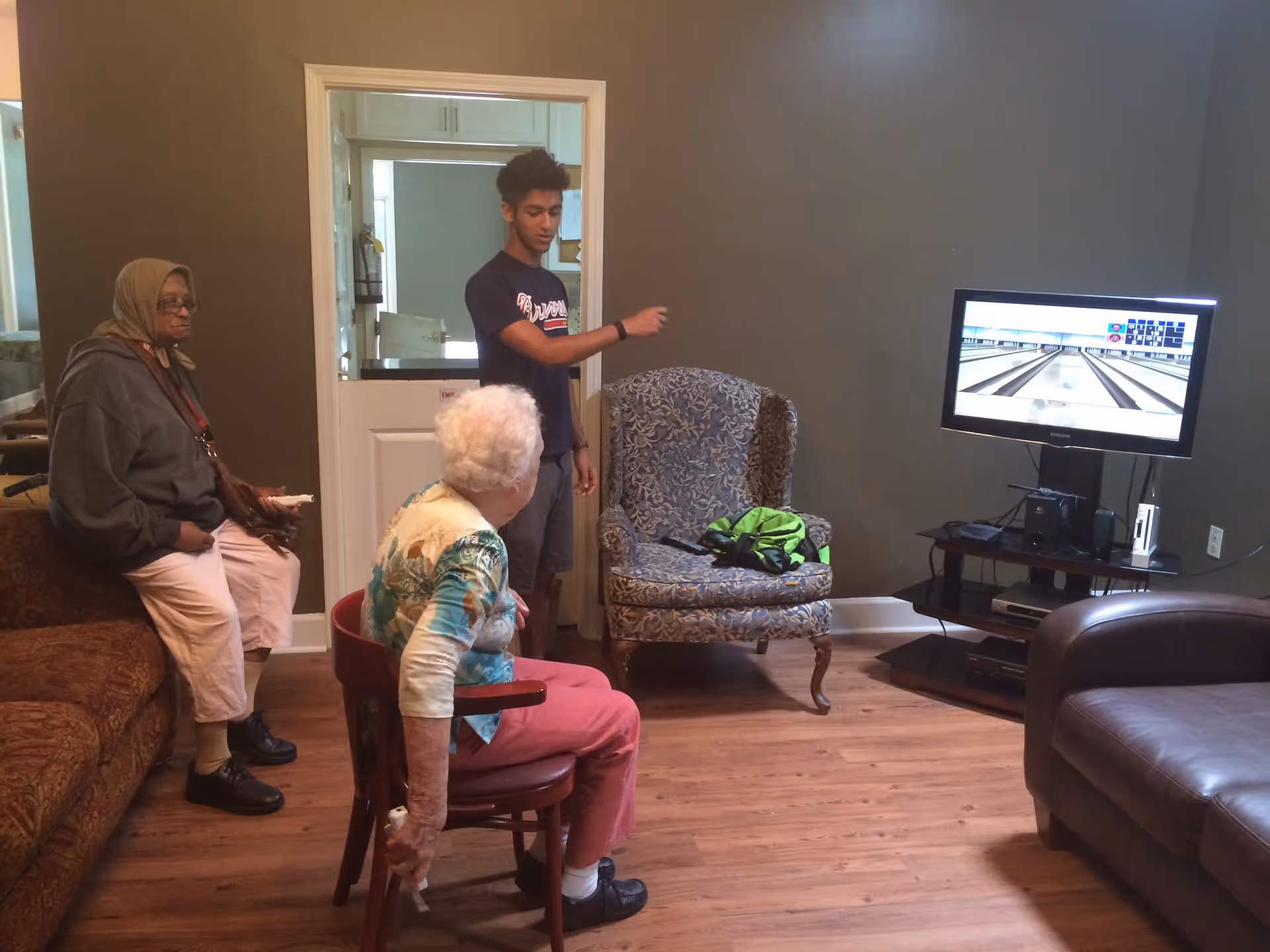 Two elderly women and a young man in a living room watching a TV bowling game.