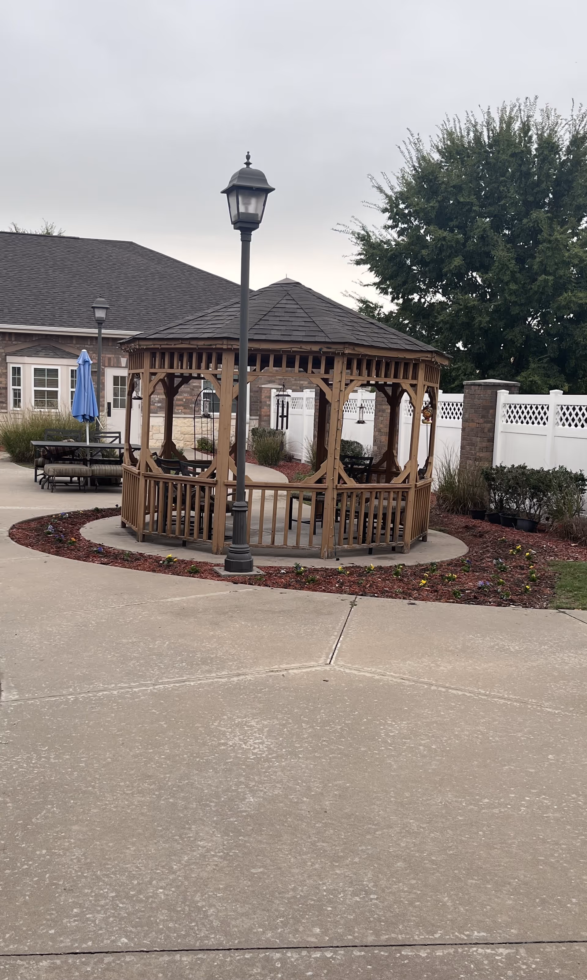 Outdoor area with a wooden gazebo surrounded by a circular flower bed with mulch and small plants. There is a lamppost in front of the gazebo, a white fence in the background, and a building with windows and a patio umbrella to the left.