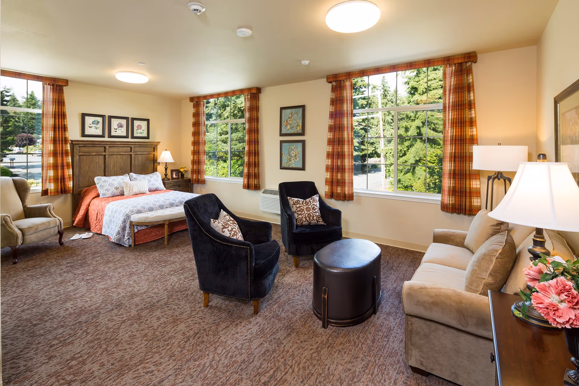 A spacious, well-lit room with a bed featuring a wooden headboard, two black armchairs with patterned pillows, a beige sofa, a small round ottoman, and large windows with plaid curtains showing green trees outside. The room has carpeted flooring, framed floral artwork on the walls, and table lamps providing warm lighting.