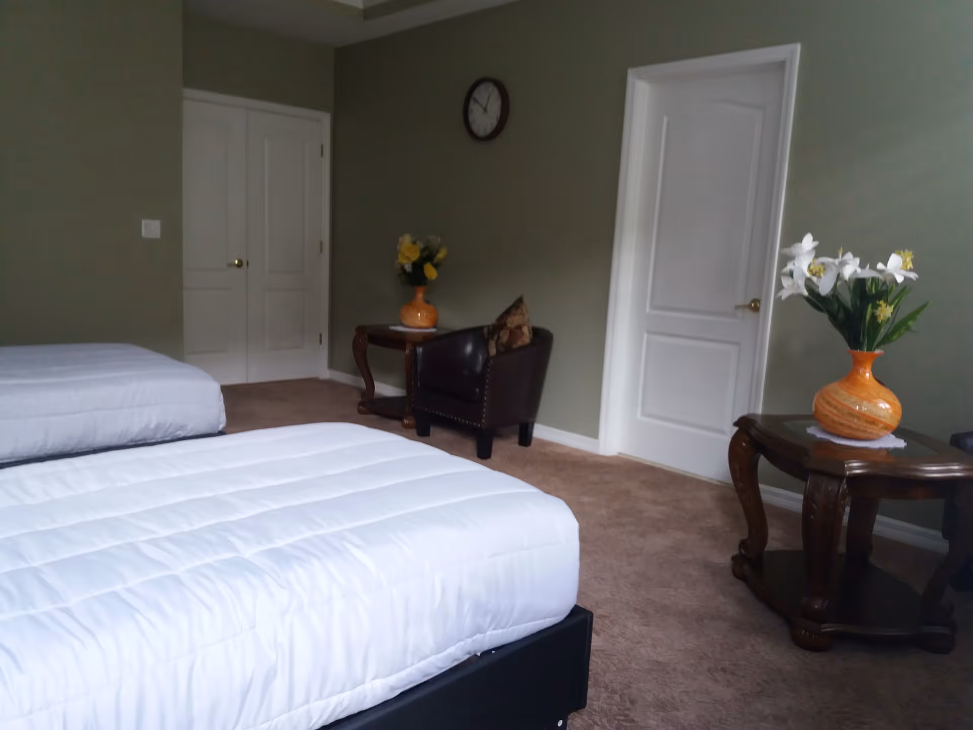 A bedroom with two single beds covered in white bedding, a brown leather armchair with a floral cushion, two wooden side tables each holding a vase with flowers, a wall clock, and two white doors against a green wall.